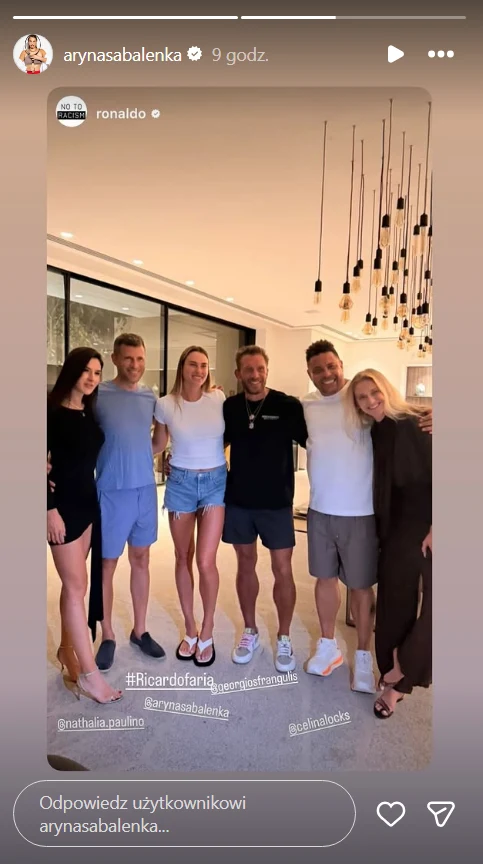 Grupa sześciu osób pozuje razem w nowoczesnym, jasnym wnętrzu, ubrane w swobodne letnie ubrania, stłoczone blisko siebie, wszyscy uśmiechnięci, w tle designerskie lampy i duże okna.