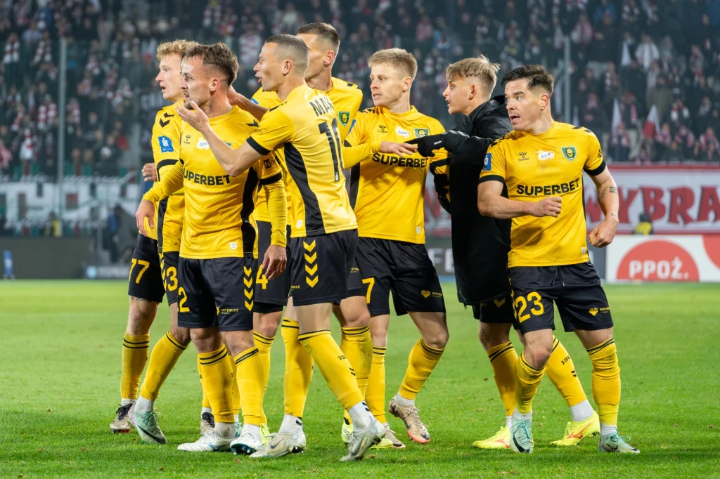 Grupa piłkarzy w żółtych strojach drużyny piłkarskiej celebruje wspólnie sukces na boisku, wyrażając radość i emocje po zdobyciu bramki lub zwycięstwie, w tle zapełnione trybuny stadionu.