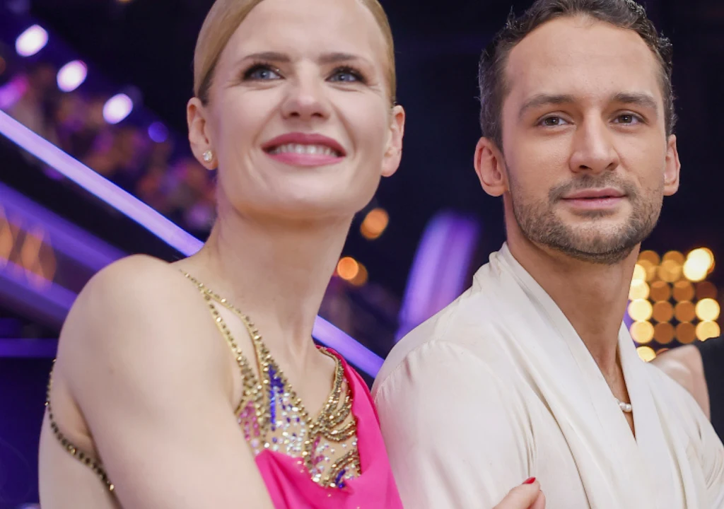 Wiktoria Gorodecka, Kamil Kuroczko Uśmiechnięta kobieta w różowej sukni trzymająca się za ramię z mężczyzną w jasnej koszuli, oboje zwróceni w stronę aparatu, widoczne kolorowe światła i rozmyte tło sugerujące atmosferę wydarzenia telewizyjnego lub gali.