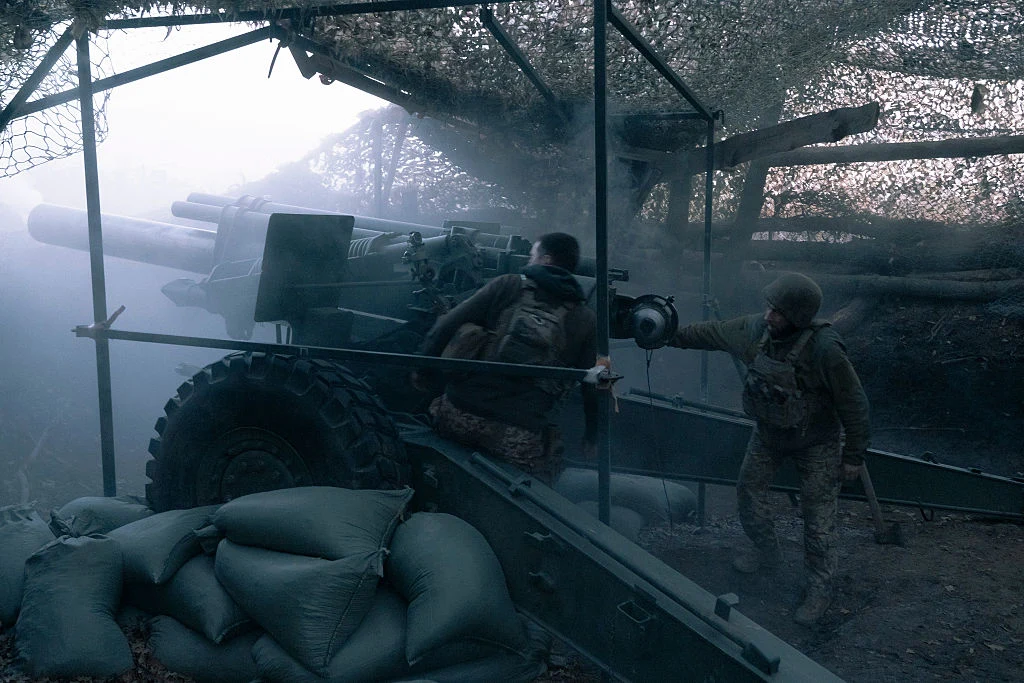 Ukraińscy żołnierze w okolicach Pokrowska Dwóch żołnierzy obsługuje ciężką armatę polową w pozycji bojowej, otoczoną workami z piaskiem, przykrytą siatką maskującą. W tle widać dym oraz zalesiony teren, atmosfera jest napięta i pełna skupienia.