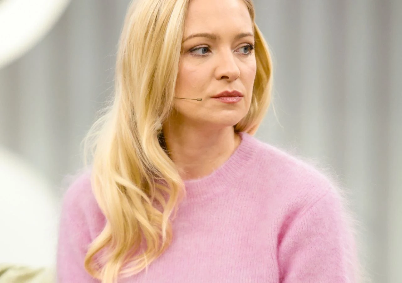 Barbara Kurdej-Szatan Kobieta o długich blond włosach, ubrana w różowy sweter, siedząca w pomieszczeniu o jasnym tle, z mikrofonem przy twarzy, patrzy w bok z neutralnym wyrazem twarzy.
