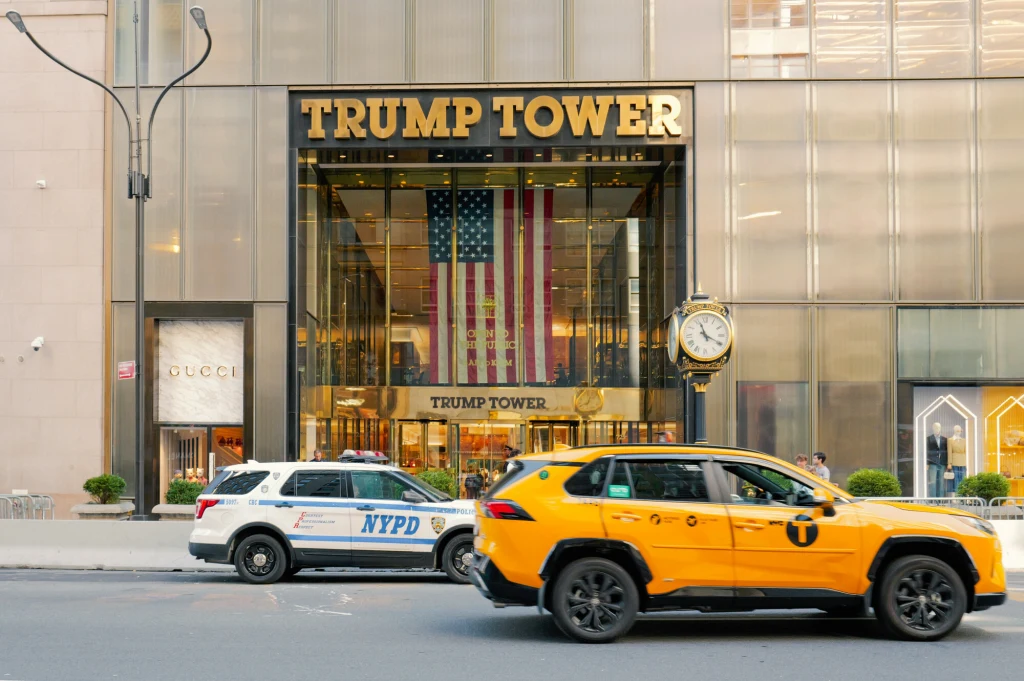 Wejście do Trump Tower, nad drzwiami duży napis z nazwą budynku oraz ogromna flaga Stanów Zjednoczonych wewnątrz, przed budynkiem żółta taksówka i radiowóz NYPD, po prawej ozdobny zegar uliczny.