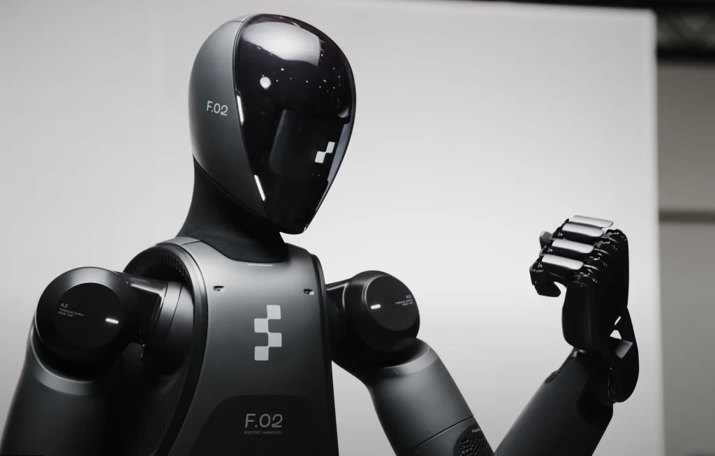 Czarny humanoidalny robot z nowoczesną, połyskującą powierzchnią, unosi rękę w geście prezentującym zaawansowaną dłoń mechaniczną, futurystyczna konstrukcja na tle białej ściany.