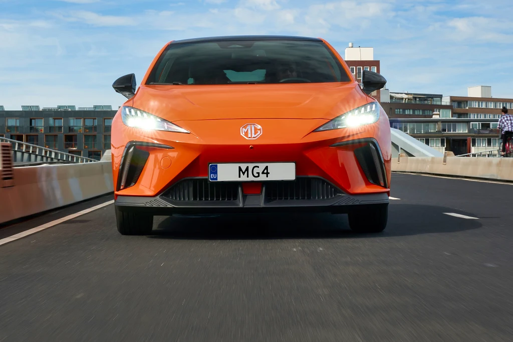 Prawie 40 proc. właścicieli MG4 EV musiało zapłacić za naprawę ponad 1 tys. funtów Pomarańczowy samochód osobowy marki MG, model MG4, sfotografowany z przodu. Jego światła są włączone, a nowoczesna linia nadwozia podkreśla dynamiczny charakter auta. W tle widoczne są budynki mieszkalne i fragment mostu.