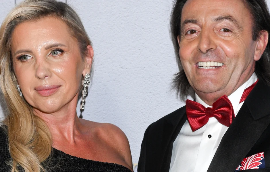 Dwoje elegancko ubranych ludzi, kobieta w czarnej sukni i mężczyzna w garniturze z czerwonym musznikiem, stoją obok siebie na tle jasnej ściany, oboje uśmiechnięci w kierunku aparatu.