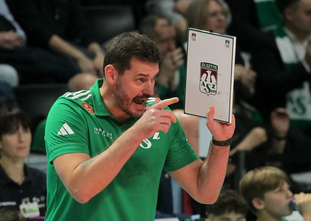 Daniel Pliński, trener Indykpolu AZS Olsztyn Trener w zielonej koszulce energicznie instruuje zespół podczas meczu, trzymając tablicę taktyczną z logo klubu sportowego. W tle widownia obserwuje przebieg wydarzeń.