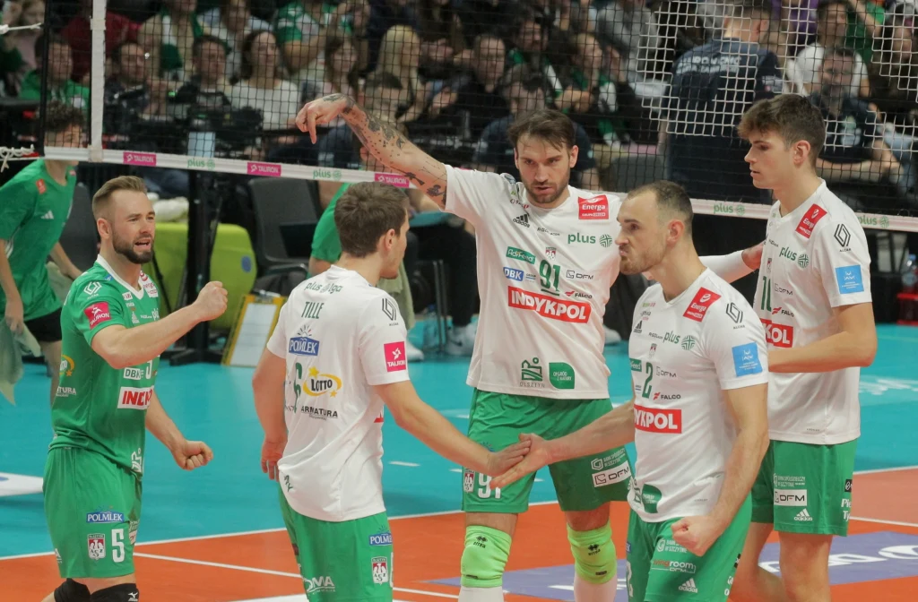 Siatkarze Indykpolu AZS Olsztyn Grupa siatkarzy w białych koszulkach podczas meczu na hali sportowej, wzajemnie motywujących się na boisku, z widocznymi logotypami sponsorów na strojach, w tle liczna publiczność za siatką.