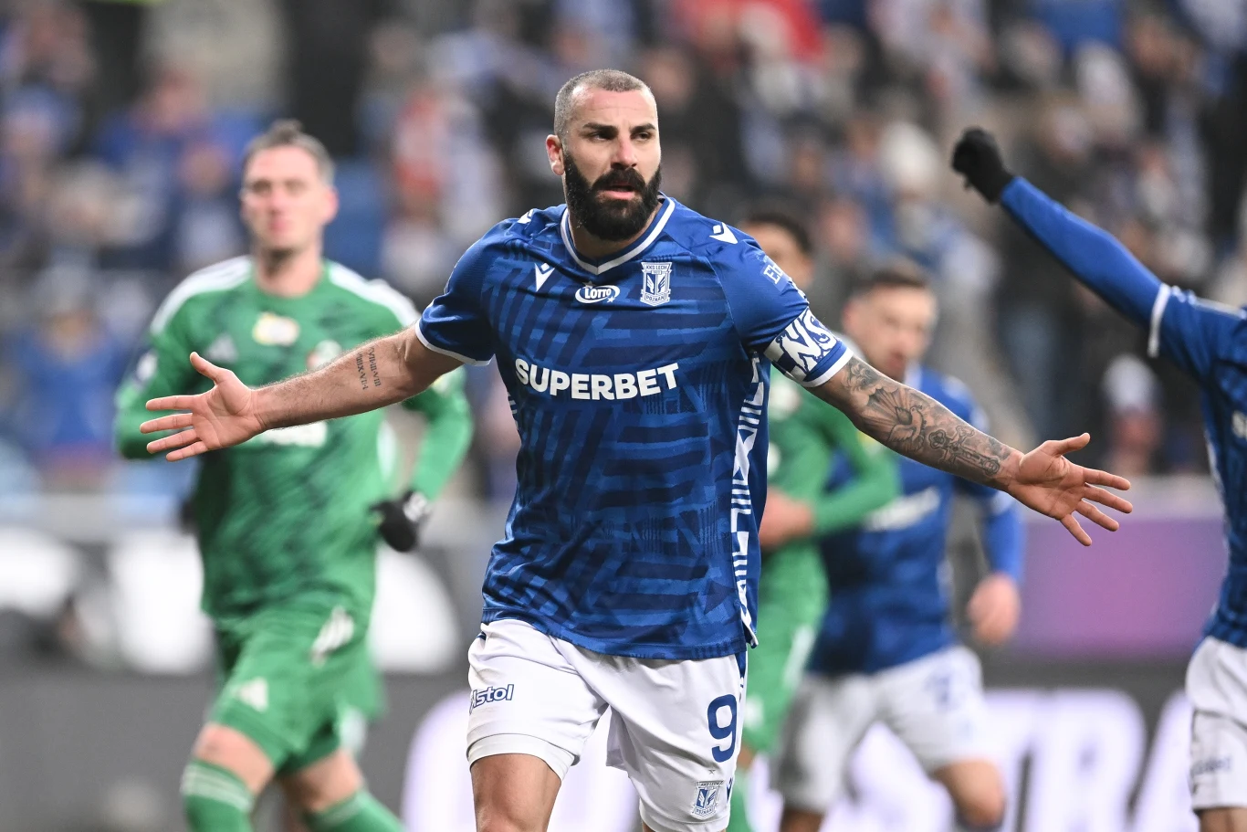 Piłkarz w niebieskim stroju drużyny Lech Poznań biegnie z szeroko rozłożonymi ramionami po zdobyciu gola, w tle widoczni zawodnicy drużyny przeciwnej w zielonych strojach oraz rozmyte trybuny stadionu.