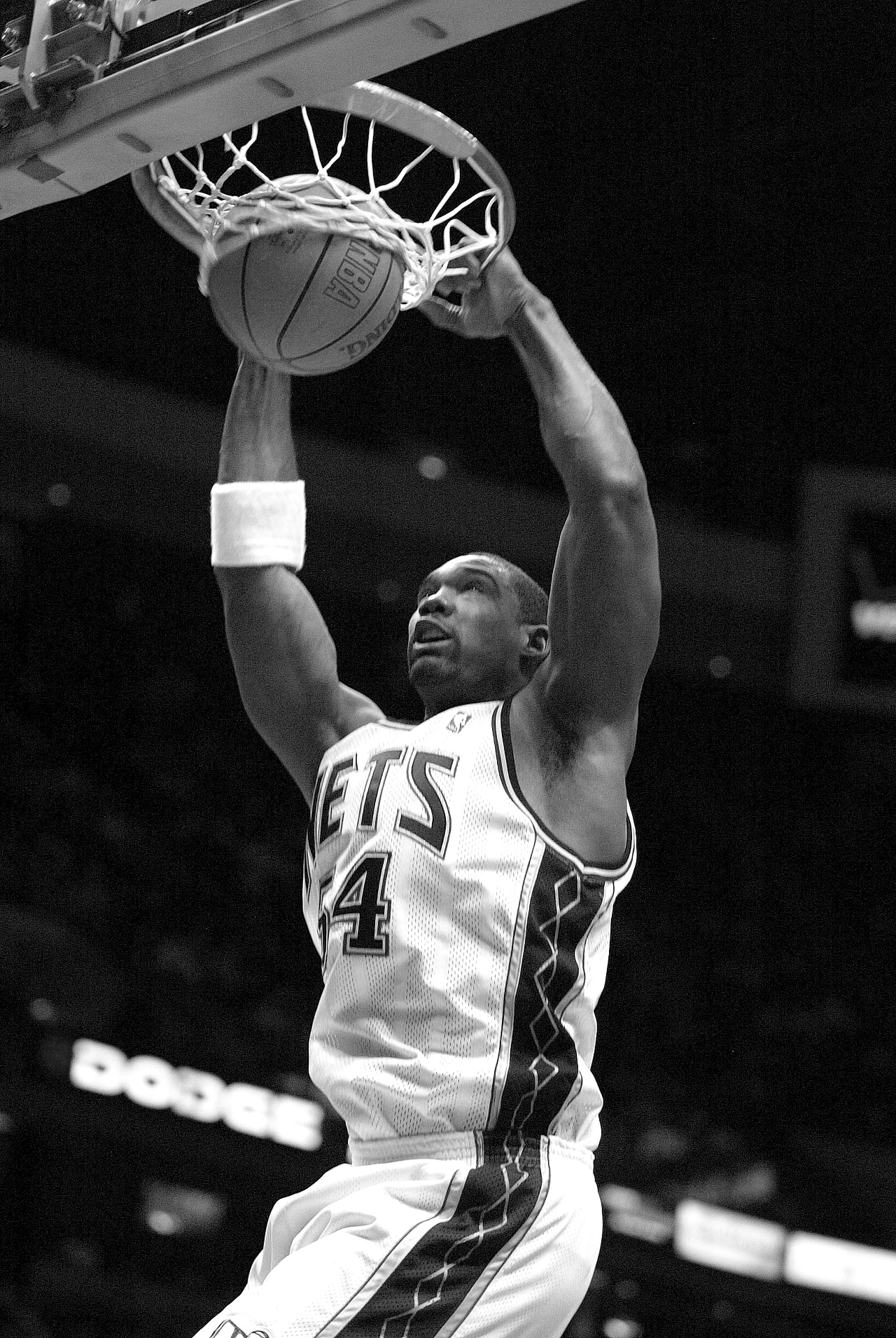 Rodney Rogers Koszykarz drużyny Nets wykonuje efektowny wsad podczas meczu, trzymając się obu rękami obręczy. Widoczna intensywność akcji oraz sportowa determinacja zawodnika. Tło lekko rozmazane, podkreślające dynamikę ruchu.