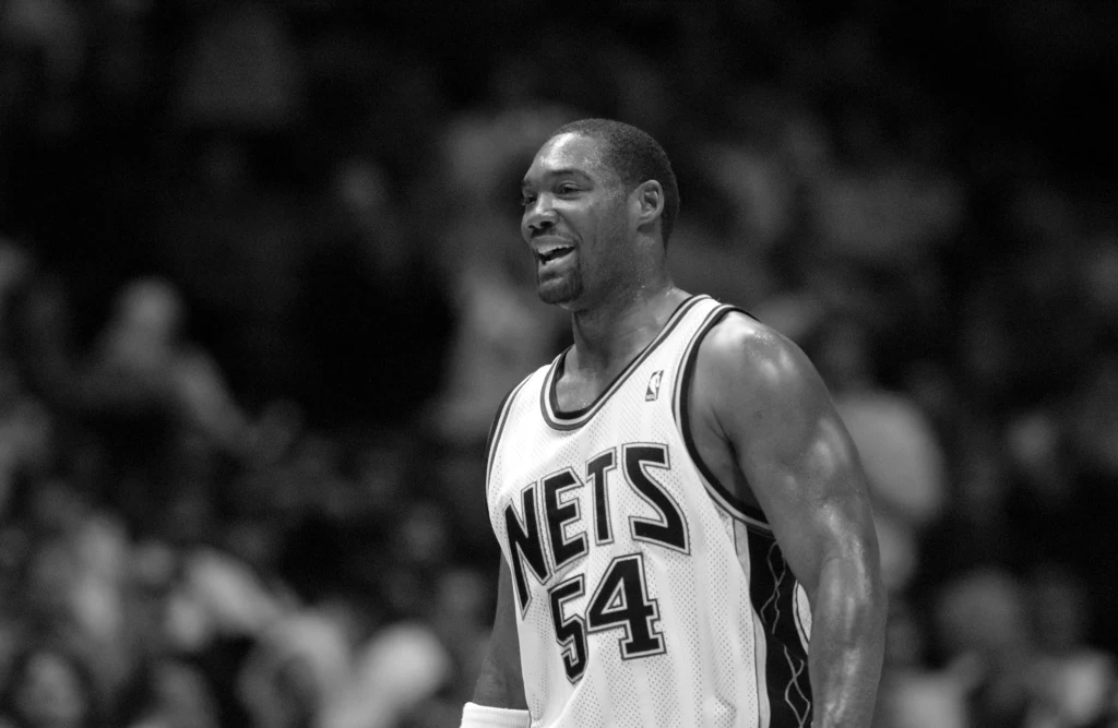 Rodney Rogers Mężczyzna w koszulce drużyny New Jersey Nets z numerem 54 na boisku koszykarskim, uśmiechnięty w trakcie gry, w tle zamazana publiczność.