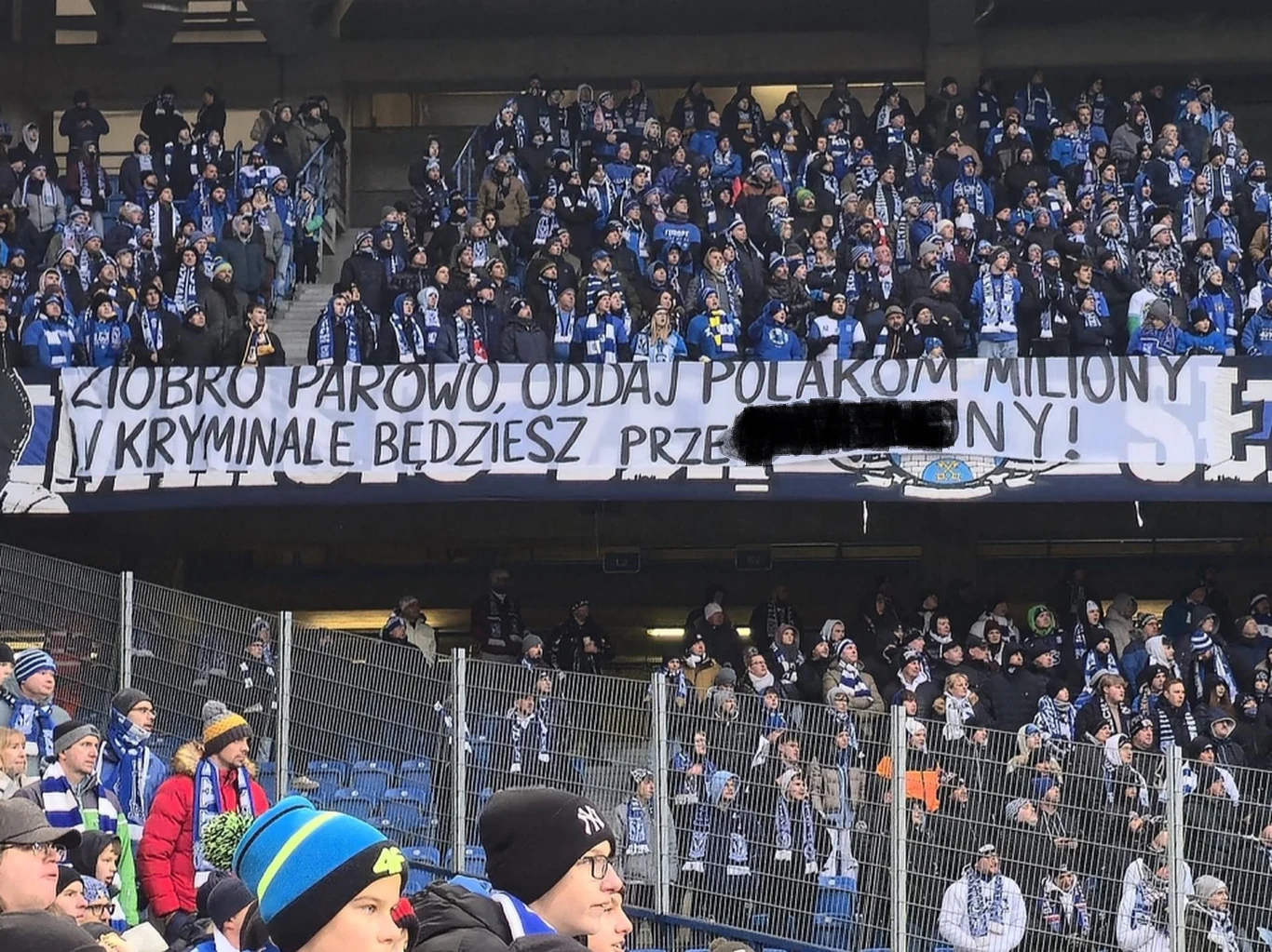 Skandaliczny transparent na meczu Lecha Poznań Tłum kibiców na stadionie trzyma rozwinięty transparent z obraźliwym i politycznie nacechowanym hasłem skierowanym przeciwko konkretnej osobie publicznej, widoczne elementy piłkarskiej oprawy oraz barwy klubowe.
