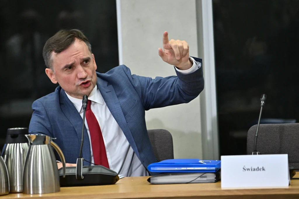 Zbigniew Ziobro Mężczyzna w garniturze i czerwonym krawacie gestykuluje palcem w górę siedząc przy stole z mikrofonem, metalowym dzbankiem i niebieskimi teczkami. Obok niego znajduje się tabliczka z napisem 'Świadek'.