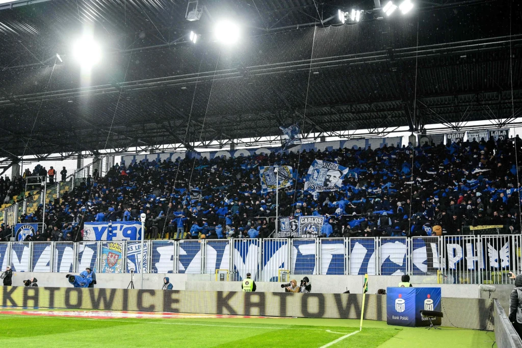 Kibice Lecha Poznań Tłum kibiców ubranych na niebiesko wypełniający trybuny stadionu, trzymający flagi i transparenty klubowe, na barierce długi transparent z napisem, w tle zadaszenie trybuny i oświetlenie stadionowe