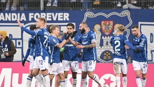 Grupa piłkarzy w niebieskich strojach celebruje zdobycie bramki na murawie stadionu, w tle widoczni kibice oraz herb klubu.