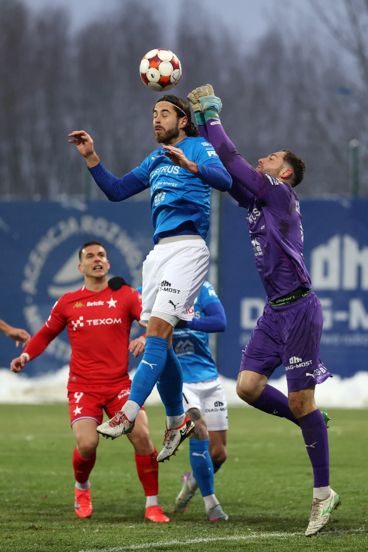 Piłkarz w niebieskim stroju wyskakuje do główki z piłką, podczas gdy bramkarz w fioletowym stroju próbuje złapać piłkę w powietrzu, w tle widoczni są kolejni zawodnicy w czerwonych strojach oraz rozmyte trybuny.