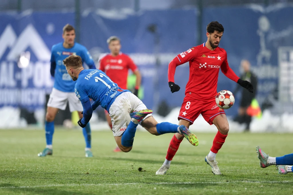 Dwóch piłkarzy w akcji na boisku, jeden w czerwonym stroju prowadzi piłkę, drugi w niebieskim wykonuje dynamiczny wślizg, w tle widoczni pozostali zawodnicy i fragment trybun.