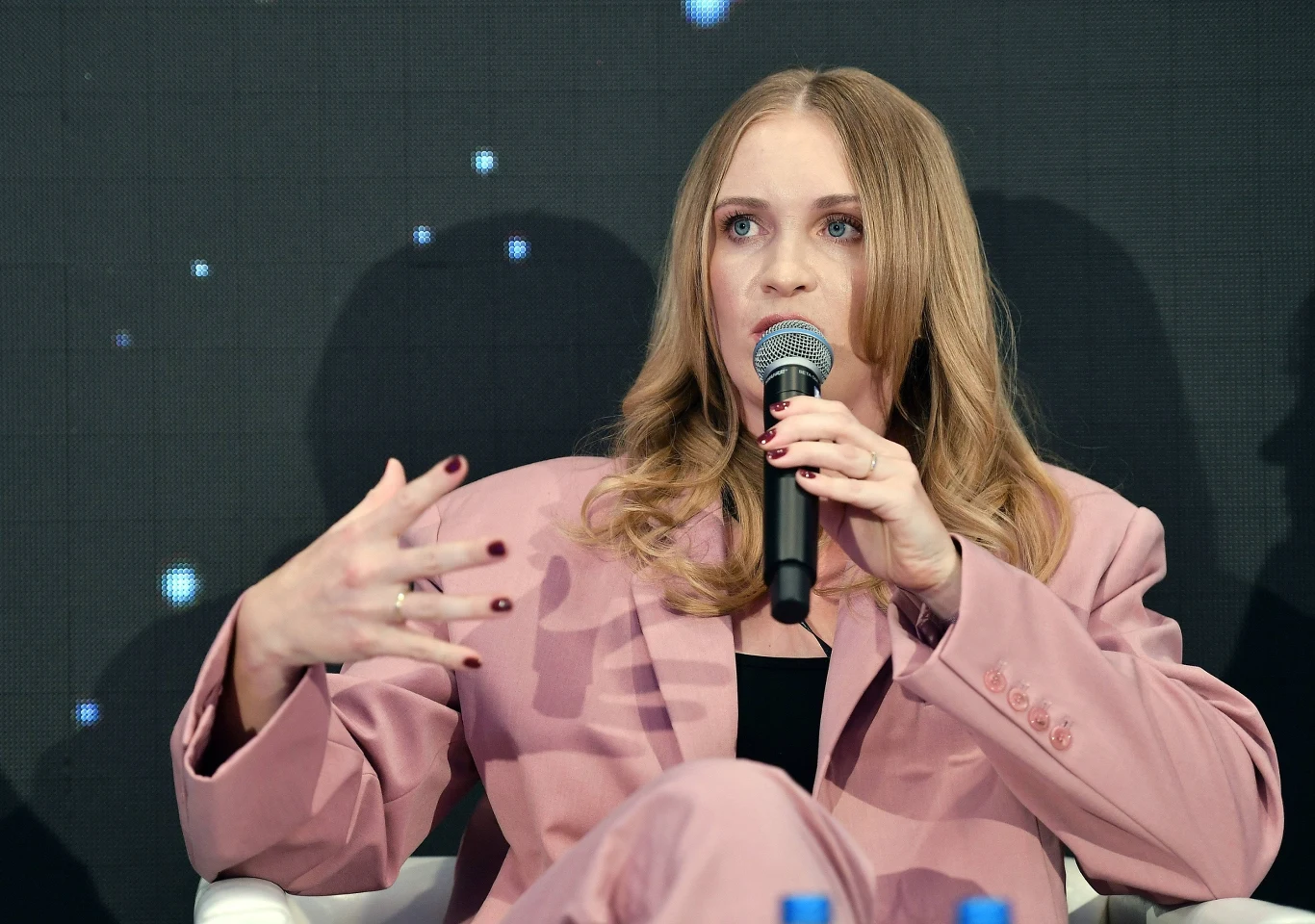 Kobieta w różowym garniturze mówi do mikrofonu, wykonując gest ręką, siedząc na tle ciemnej ścianki z jasnymi punktami świetlnymi.