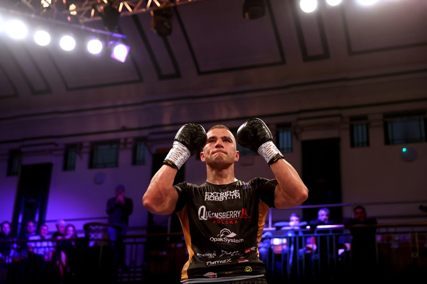 Michał Soczyński Bokser w ringu unosi rękawice bokserskie w geście triumfu, na tle rozświetlonej hali z publicznością widoczną w tle.