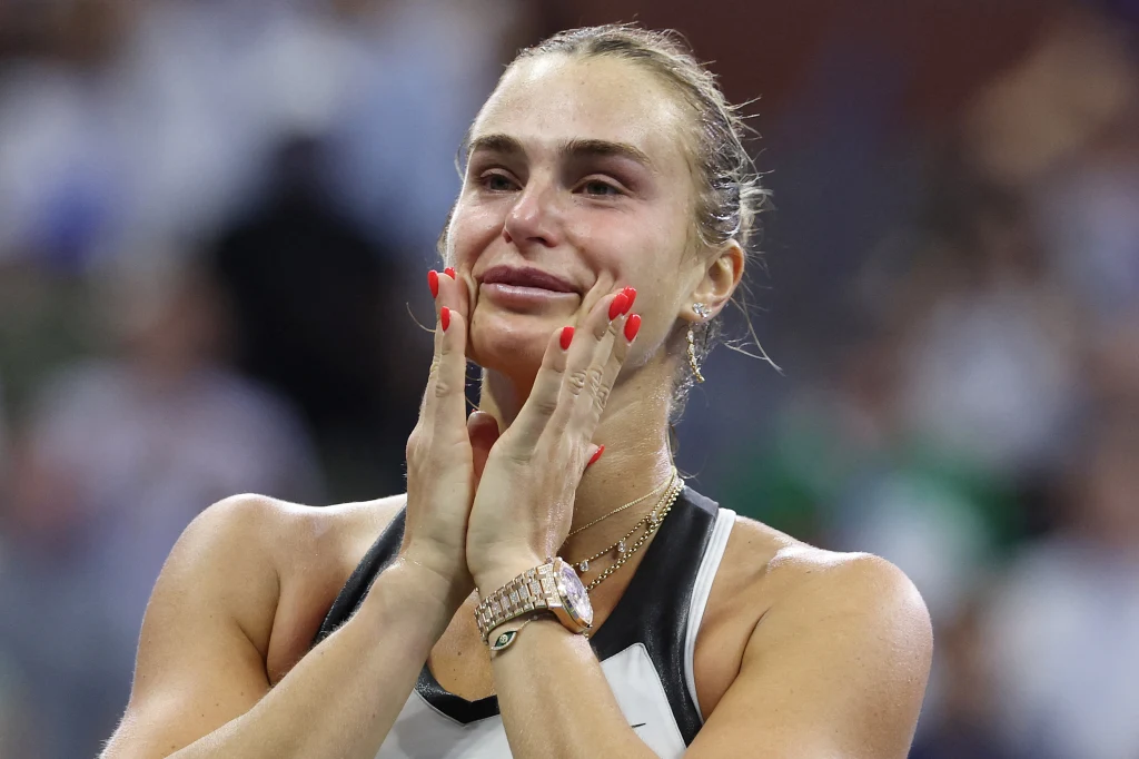 Aryna Sabalenka nie kryła wzruszenia po wygraniu US Open 2025 Kobieta ze wzruszoną miną trzyma dłońmi twarz, na jej twarzy widać łzy wzruszenia. Ubrana jest w sportowy strój i złote naszyjniki, a na dłoniach ma czerwony lakier do paznokci. W tle rozmyte sylwetki innych osób.