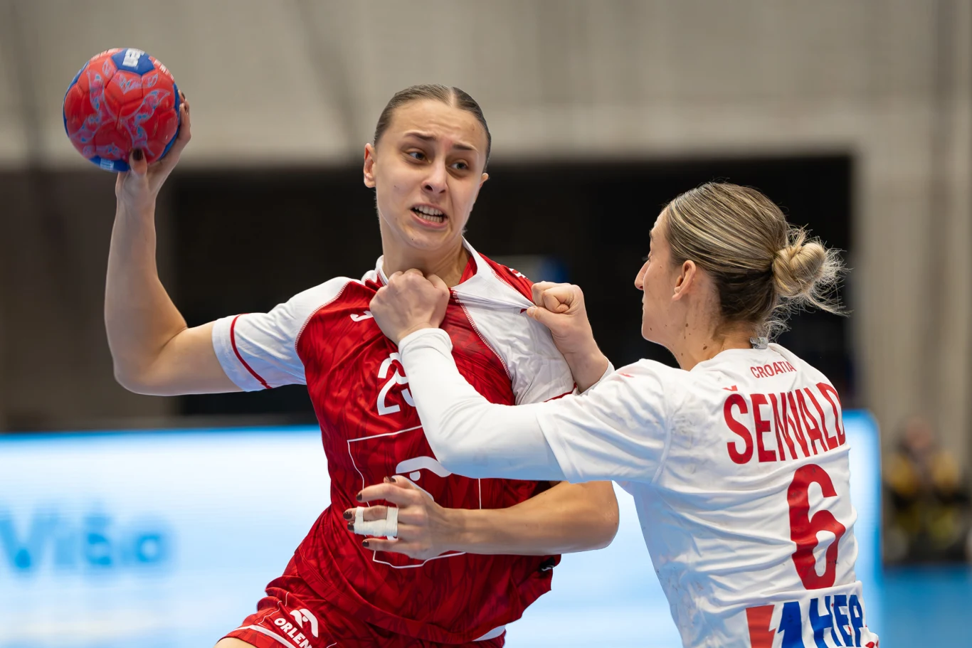 Zawodniczka w czerwonym stroju trzyma piłkę ręczną i próbuje przedrzeć się przez obronę rywalki w białej koszulce, która chwyta ją za koszulkę, próbując zatrzymać akcję.