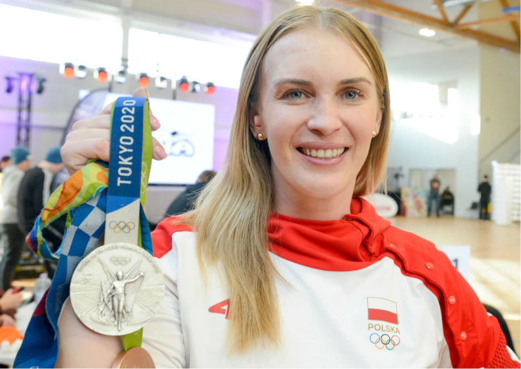 Kobieta w sportowym stroju z orzełkiem i napisem Polska trzyma w ręce srebrny medal olimpijski oznaczony napisem Tokyo 2020, uśmiecha się do kamery na tle jasnego pomieszczenia z widocznymi elementami hali sportowej.
