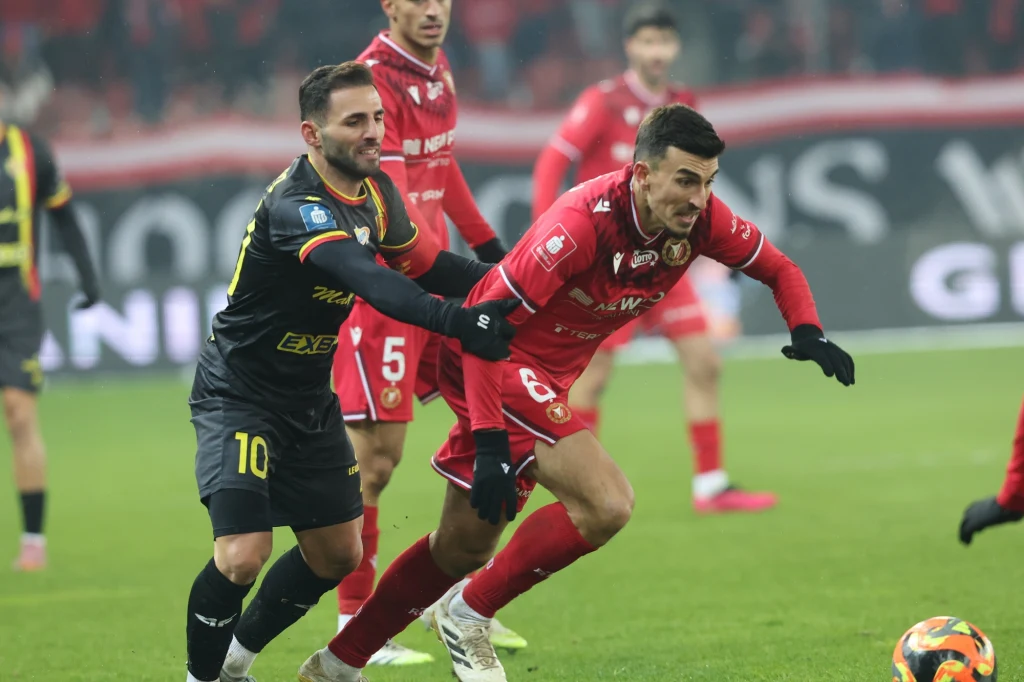 Juljan Shehu (z prawej) znalazł drogę do bramki rywala, ale dla Widzewa było to tylko trafienie honorowe Dwóch piłkarzy w trakcie dynamicznej akcji na boisku, jeden w czarnej koszulce z numerem 10 próbuje przejąć piłkę od zawodnika w czerwonym stroju, tło rozmazane z widocznymi innymi graczami.