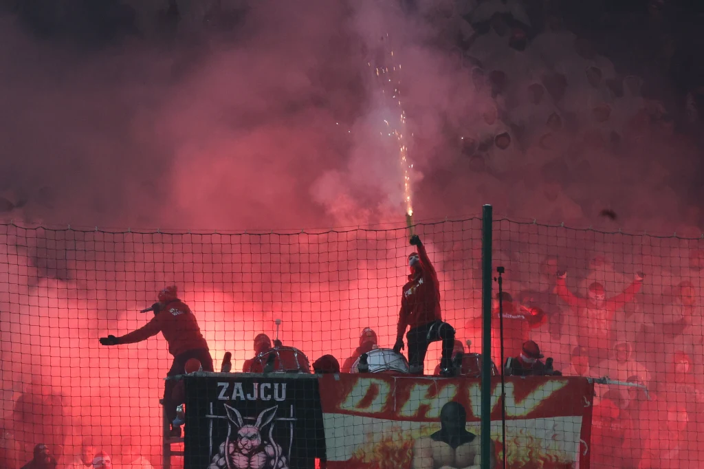 Kibice Widzewa liczyli tego dnia na znacznie więcej Grupa kibiców za ogrodzeniem stadionowym wznosząca okrzyki, jeden z nich odpala racę, wokół unosi się gęsty czerwony dym, w tle widoczne transparenty i wyraźna atmosfera napięcia.