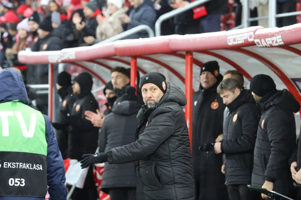 Trener Igor Jovicević na razie ma kłopot... z obietnicą, którą złożył krótko po podpisaniu kontraktu z RTS Ławka rezerwowych drużyny piłkarskiej podczas meczu, trener wydaje polecenia, wokół stoją inni członkowie sztabu oraz zawodnicy w zimowych kurtkach, w tle widać kibiców na trybunach.
