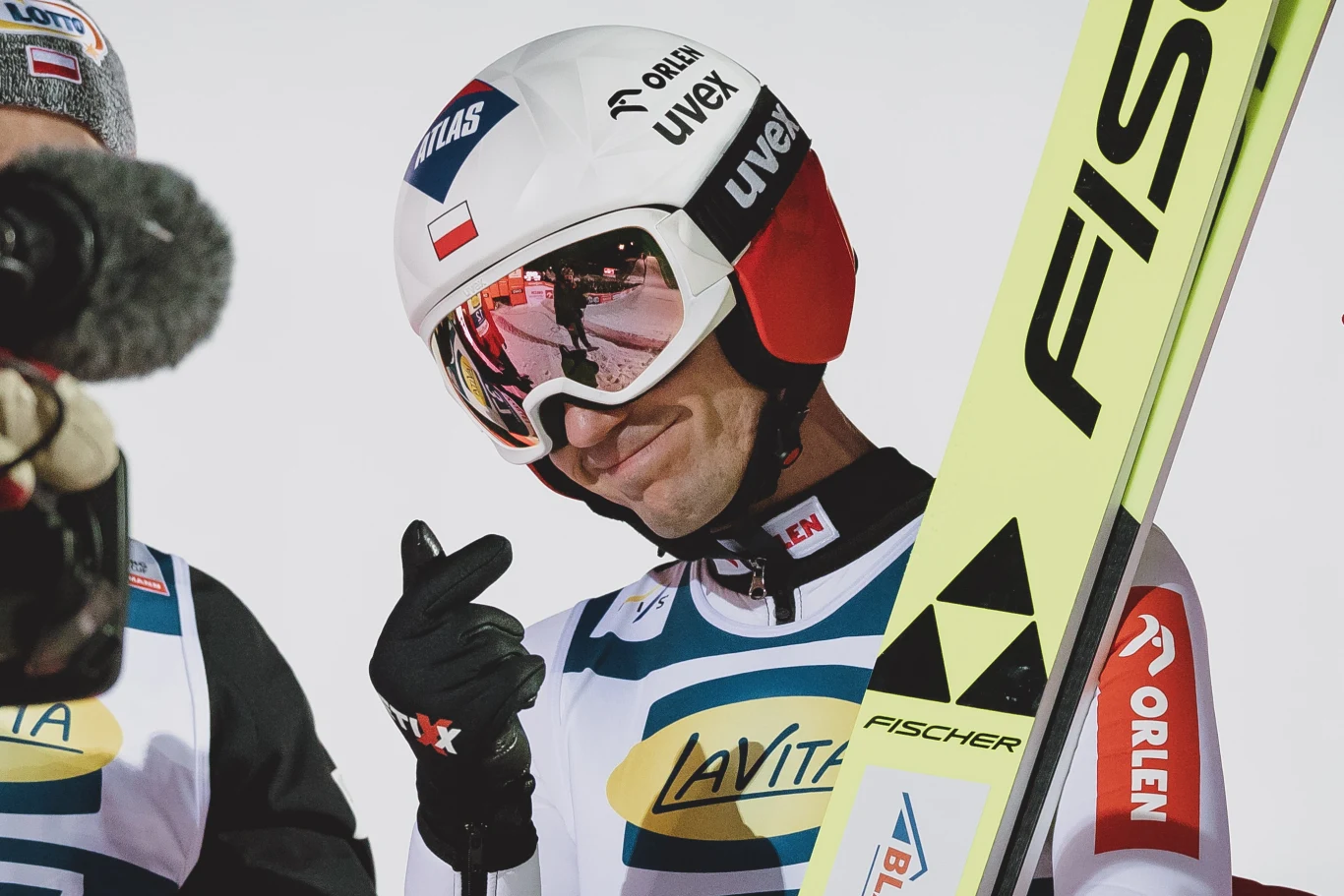 Kamil Stoch Zawodnik w stroju narciarskim trzyma narty, na kasku widoczne logo sponsorów oraz polska flaga, pokazuje gest dłonią w kierunku obiektywu, obok znajduje się inna osoba z kamerą.