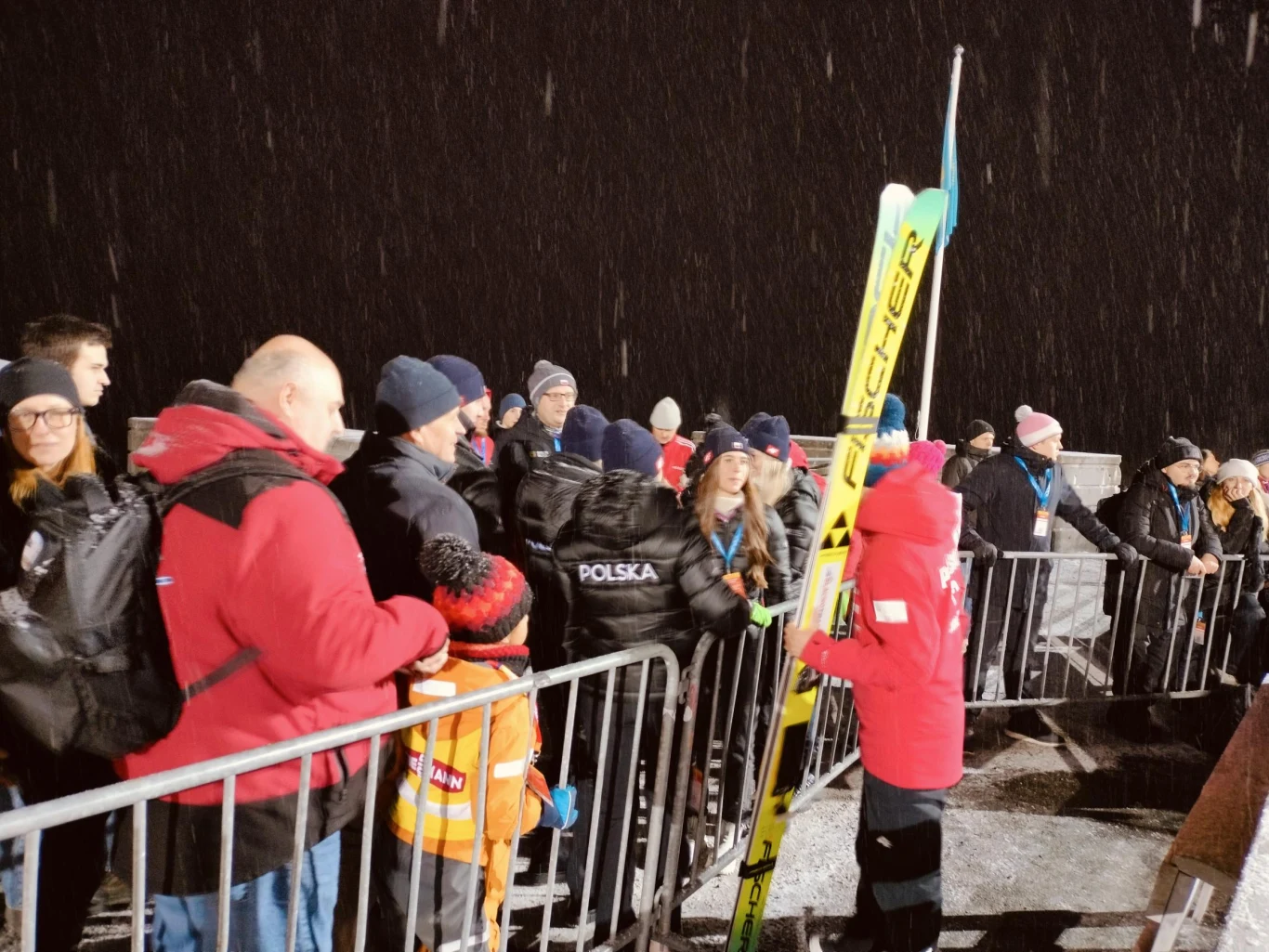 Kamil Stoch z kibicami i reprezentacją Polski biathlonistek w Lillehammer Tłum ludzi ubranych w zimowe kurtki stoi za barierkami, niektórzy mają na głowach czapki. Na pierwszym planie osoba w czerwonym stroju trzyma narty skokowe z logo Fischer. Wieczorna sceneria i padający śnieg podkreślają atmosferę wydarzenia sportowego.