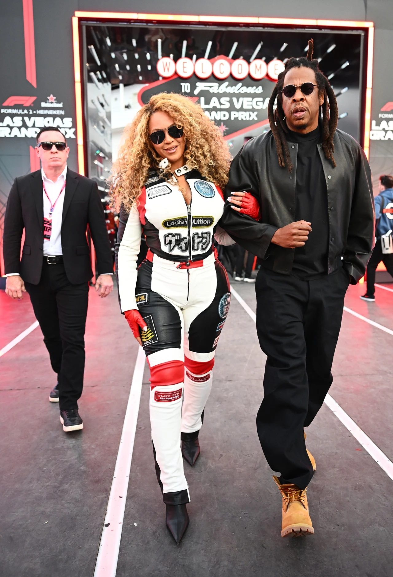 Dwoje znanych celebrytów idzie razem, trzymając się pod ramię, na tle wejścia do hali wyścigów Formuły 1 w Las Vegas. Kobieta ubrana w kombinezon wyścigowy z naszywkami i okularami przeciwsłonecznymi, mężczyzna w czarnej kurtce i dżinsach. W tle widocz...