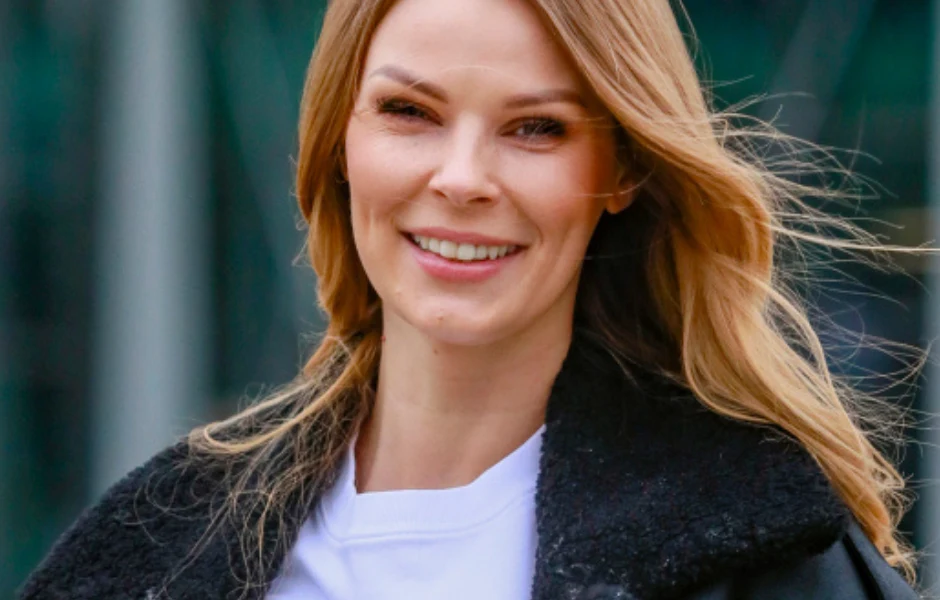 Uśmiechnięta kobieta z jasnymi włosami ubrana w białą koszulkę oraz czarną kurtkę, na tle nieostrego, miejskiego krajobrazu.