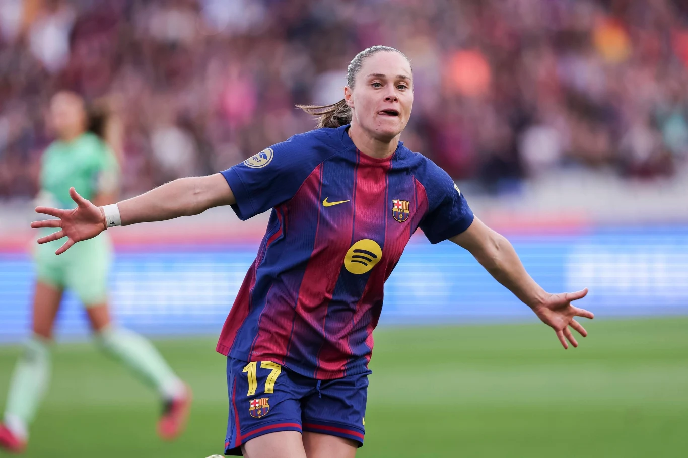 Piłkarka w stroju FC Barcelony świętuje zdobycie bramki z szeroko rozłożonymi ramionami, w tle widać rozmazane sylwetki innych zawodniczek oraz trybuny pełne kibiców.