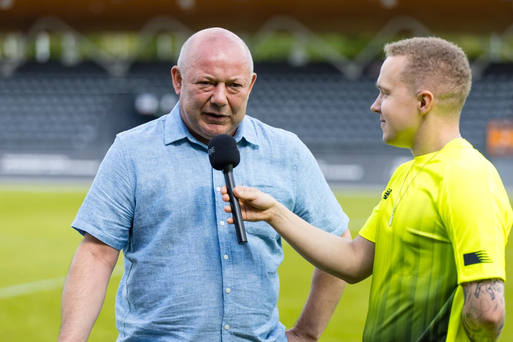Wojciech Kowalczyk w roli futbolowego eksperta Starszy mężczyzna w niebieskiej koszuli udziela wywiadu na stadionie, mikrofon trzyma młody mężczyzna w żółtej koszulce sportowej.