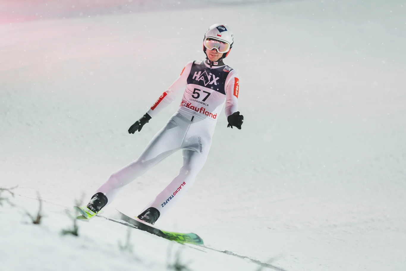 Kamil Stoch Skoczek narciarski w białym kombinezonie ląduje na skoczni, w tle widać zasypaną śniegiem ślizgawkę i opadający śnieg. Numer startowy 57 oraz logo sponsorów na stroju sportowca. Zawodnik w pełnym ekwipunku, z kaskiem i goglami.