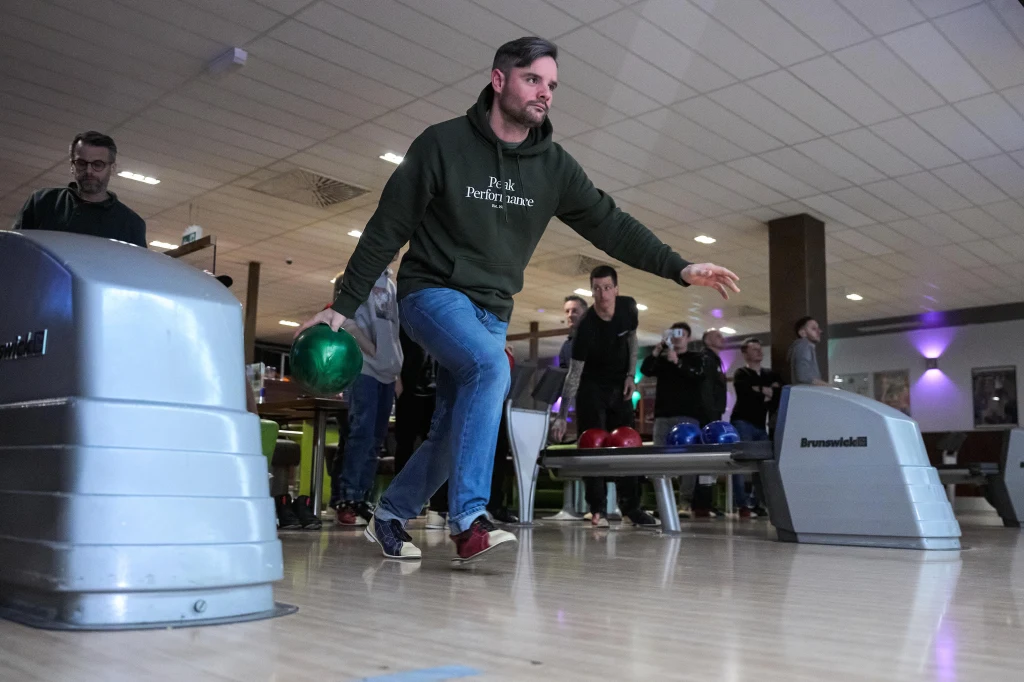 Mężczyzna w zielonej bluzie rzuca kulę do kręgli na torze w kręgielni, w tle kilku obserwujących ludzi oraz inne kolorowe kule i wyposażenie kręgielni.