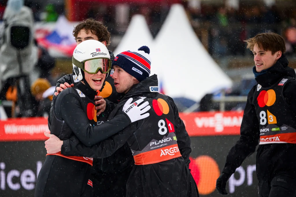 Grupa skoczków narciarskich ubrana w kombinezony z numerami startowymi, widoczne emocje i radość po zakończonym występie, zimowa sceneria, w tle rozmazane postaci oraz namioty.