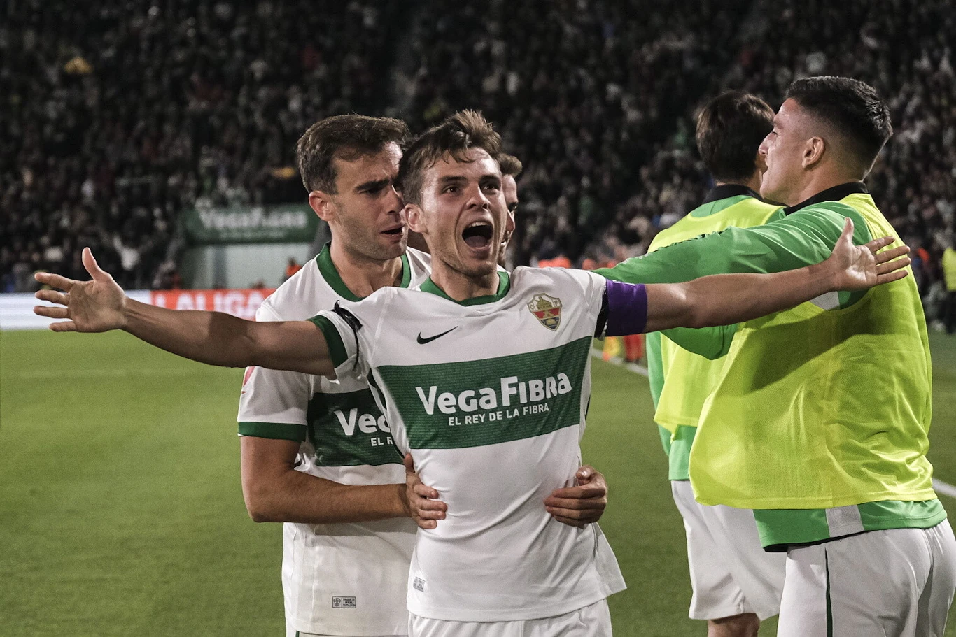 Piłkarz w białej koszulce z rozłożonymi rękami świętuje zdobycie gola, wokół niego zespołowi koledzy w podobnych strojach wyrażają radość, w tle widać trybuny pełne kibiców.