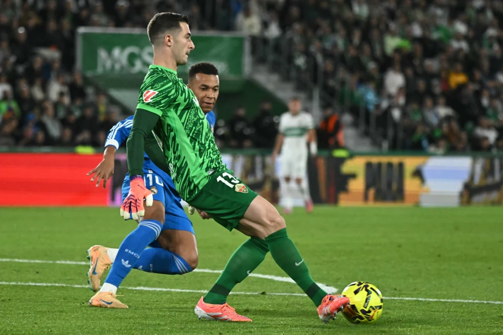 Dwóch piłkarzy walczy o piłkę na boisku podczas meczu, jeden ubrany w zielony strój wykonuje precyzyjny ruch nogą próbując przejąć piłkę, drugi zawodnik w niebieskim stroju znajduje się tuż za przeciwnikiem. W tle widać kibiców na trybunach i fragmenty...