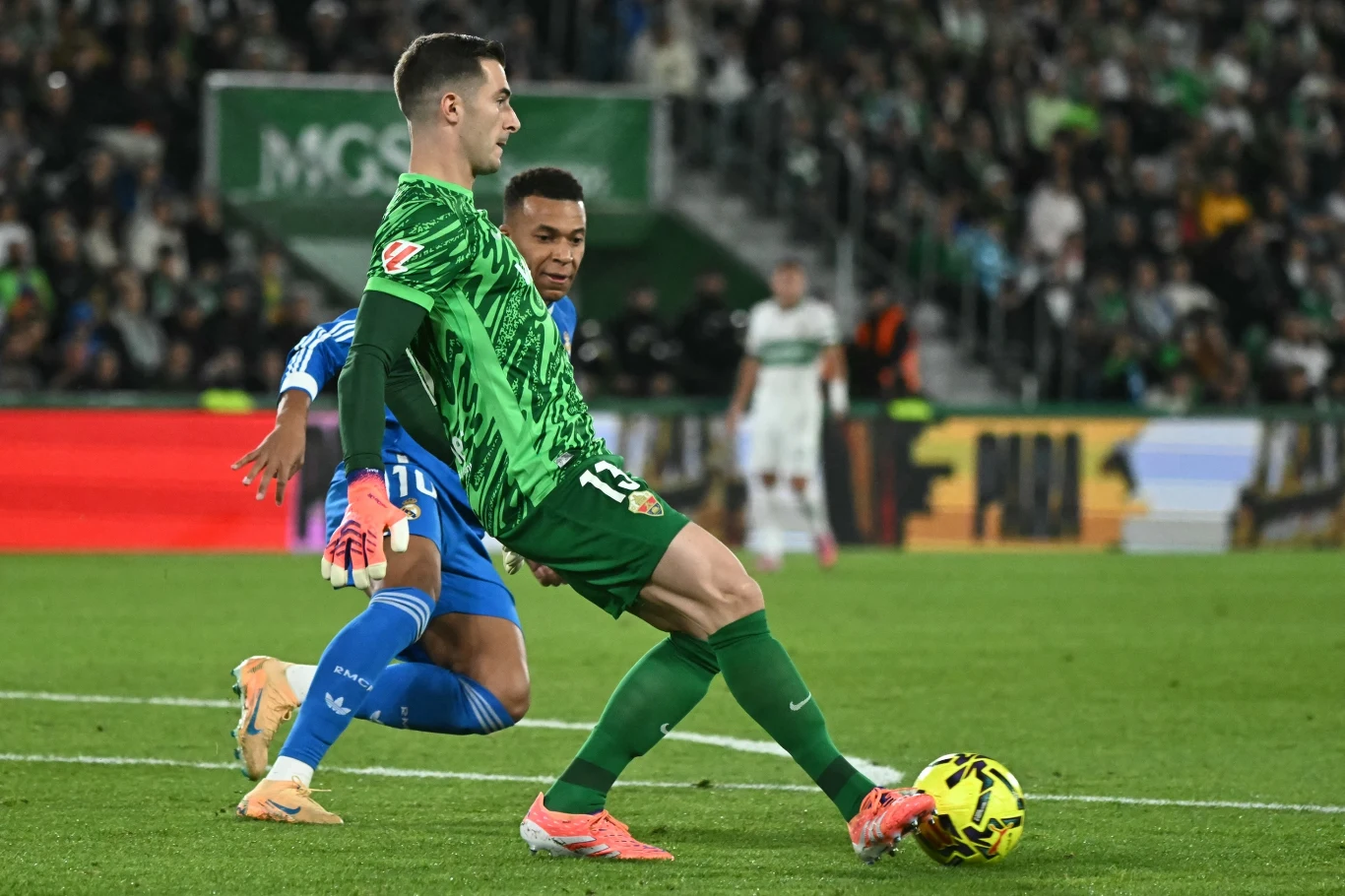 Dwóch piłkarzy walczy o piłkę na boisku podczas meczu, jeden ubrany w zielony strój wykonuje precyzyjny ruch nogą próbując przejąć piłkę, drugi zawodnik w niebieskim stroju znajduje się tuż za przeciwnikiem. W tle widać kibiców na trybunach i fragmenty...