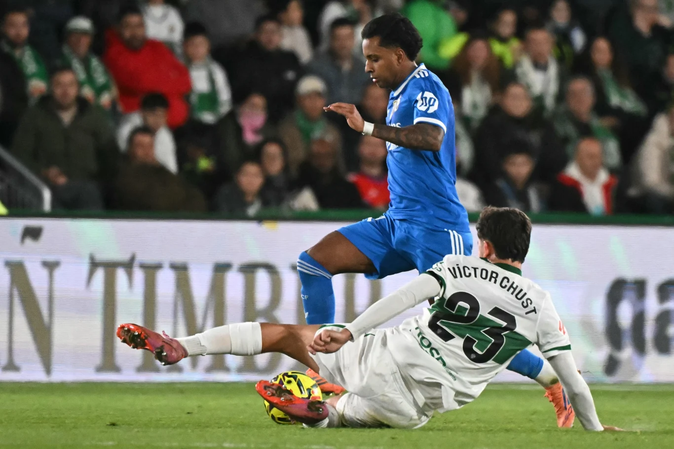 Piłkarz w niebieskim stroju unika wślizgu zawodnika w białej koszulce na tle pełnych trybun stadionu.