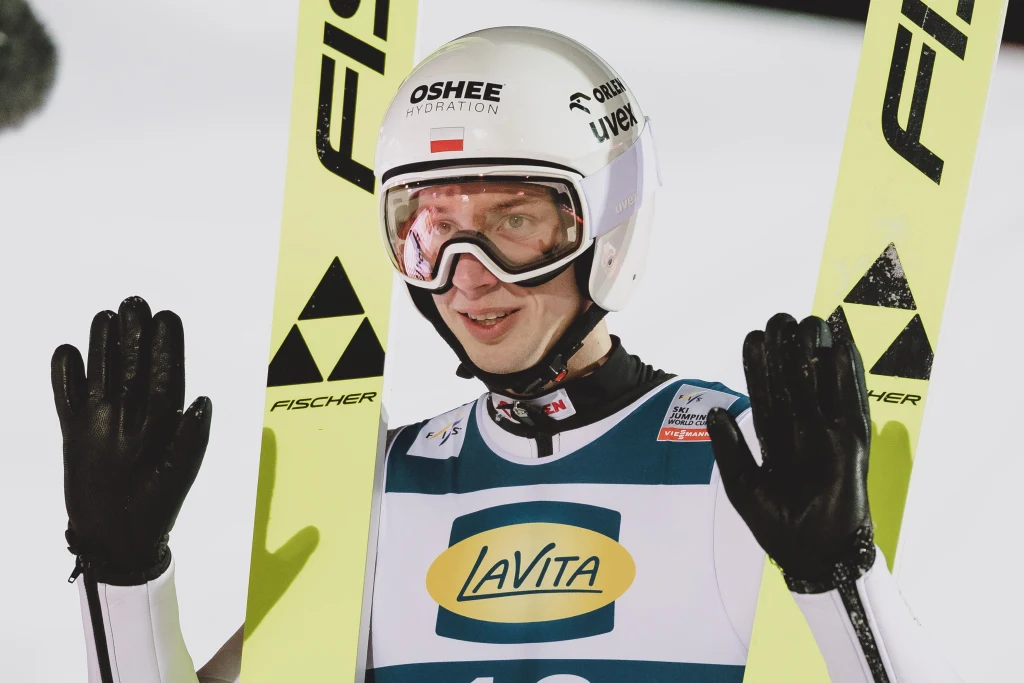 Skoczek narciarski ubrany w biały kask, gogle i kombinezon z widocznymi logotypami sponsorów trzyma w dłoniach narty. Twarz sportowca wyraża emocje, a w tle widać fragment skoczni pokrytej śniegiem.