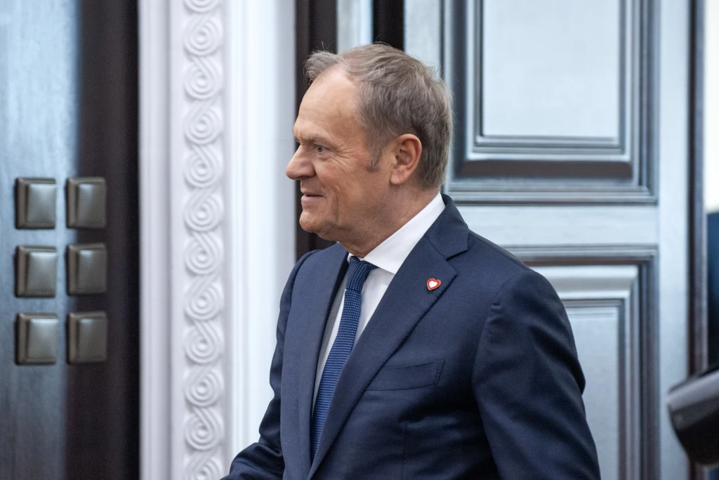 Premier Donald Tusk Mężczyzna w garniturze i krawacie stoi bokiem w eleganckim wnętrzu z ozdobną białą sztukaterią i drewnianymi drzwiami w tle. Na klapie marynarki widoczna przypinka z polskim symbolem narodowym.