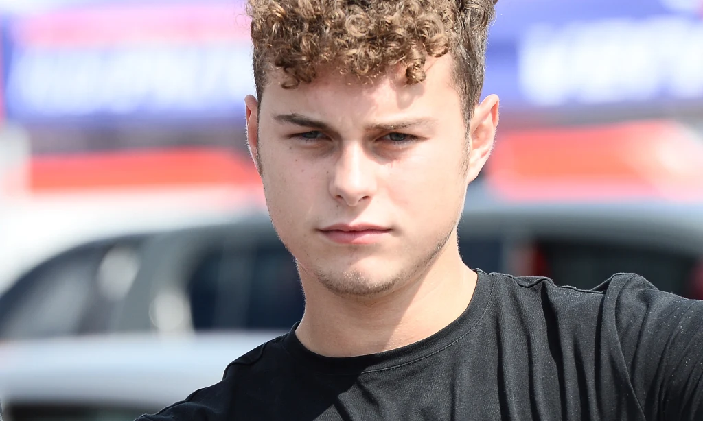 Adam Zdrójkowski Młody mężczyzna z kręconymi włosami i jasną cerą w czarnej koszulce patrzy prosto w obiektyw, na tle rozmytego krajobrazu miejskiego z samochodami i reklamami.