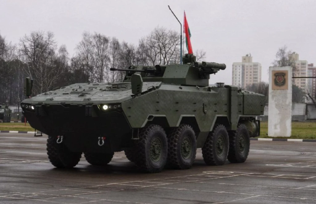 Nowoczesny pojazd opancerzony na kołach, pomalowany na ciemnozielony kolor, z wysuniętą wieżyczką z uzbrojeniem, jadący po pustym placu z flagą zamocowaną na górze. W tle znajdują się drzewa bez liści oraz kilka wysokich budynków mieszkalnych.