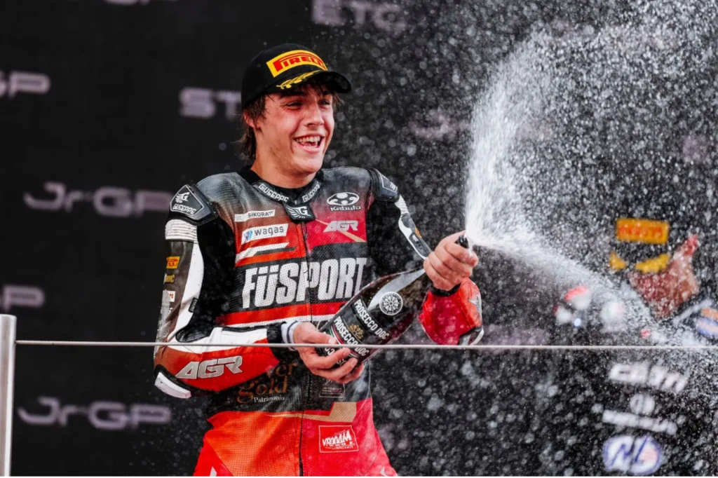 Milan Pawelec mistrzem Europy Moto2! Motocyklista w stroju sportowym i czapce rozlewa szampana podczas celebracji na podium, wyraźnie uśmiechnięty i podekscytowany, w tle ciemne tło z logotypami sponsorskimi.