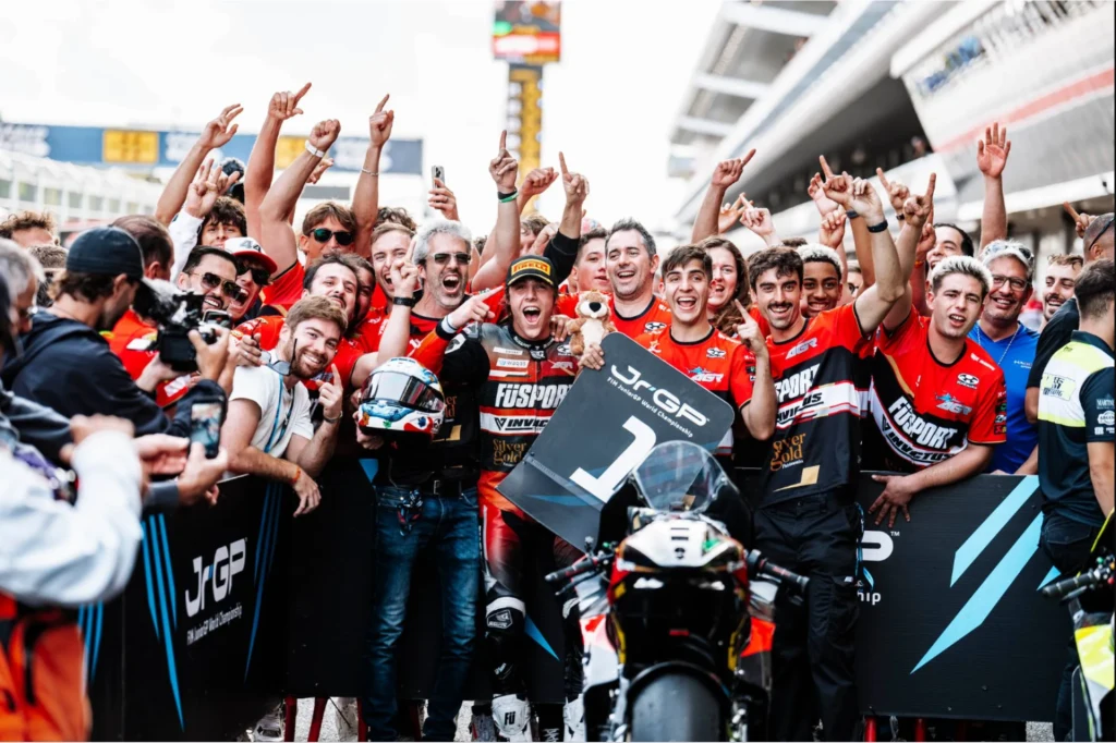 Historyczny sukces Pawelca. 18-latek został mistrzem Europy Moto2. Grupa ubranych w czerwono-czarne stroje członków zespołu wyścigowego, świętująca zwycięstwo z motocyklistą trzymającym puchar i stojącym na pierwszym miejscu z tablicą symbolizującą wygraną, motocyklem na pierwszym planie oraz tłumem podnoszącym ręce w...