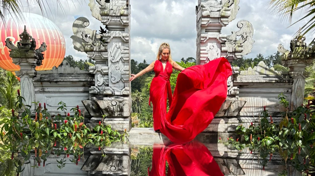 Kobieta w efektownej, długiej czerwonej sukni stoi pomiędzy ozdobnymi białymi kolumnami na tle egzotycznej roślinności, a jej odbicie widoczne jest w lśniącej powierzchni pod stopami.
