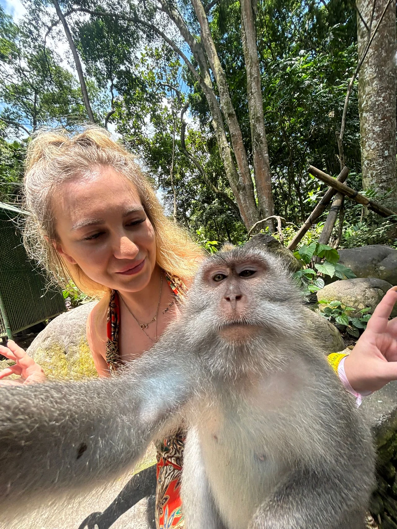 Małpa wykonująca selfie z młodą kobietą w otoczeniu zielonych drzew i naturalnej roślinności, w słoneczny dzień.