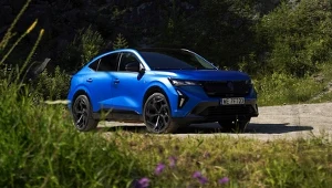 Niebieski samochód typu SUV coupe stoi na szutrowej drodze otoczonej zielenią i roślinnością, w tle widać drzewa i wzniesienia.